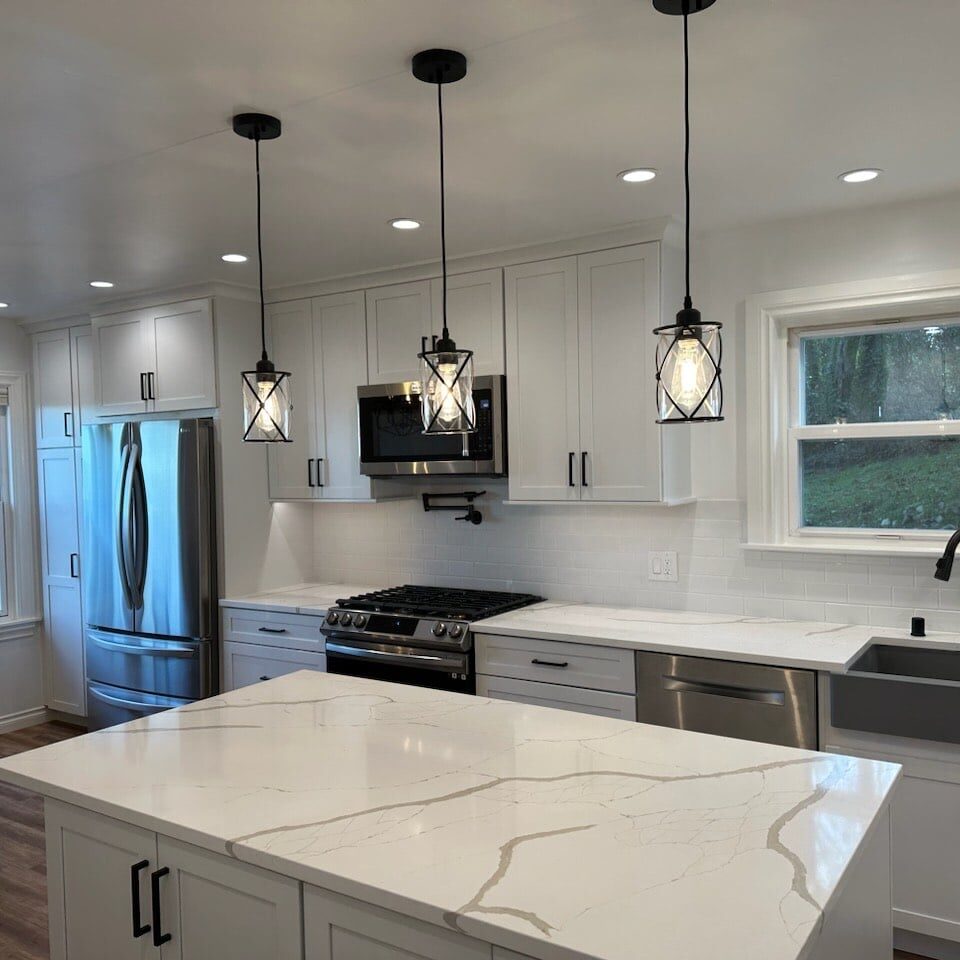 Modern kitchen with white cabinets, marble island, stainless steel appliances, three pendant lights, large sink, and windows overlooking greenery—expertly crafted by a West Seattle remodeling contractor.