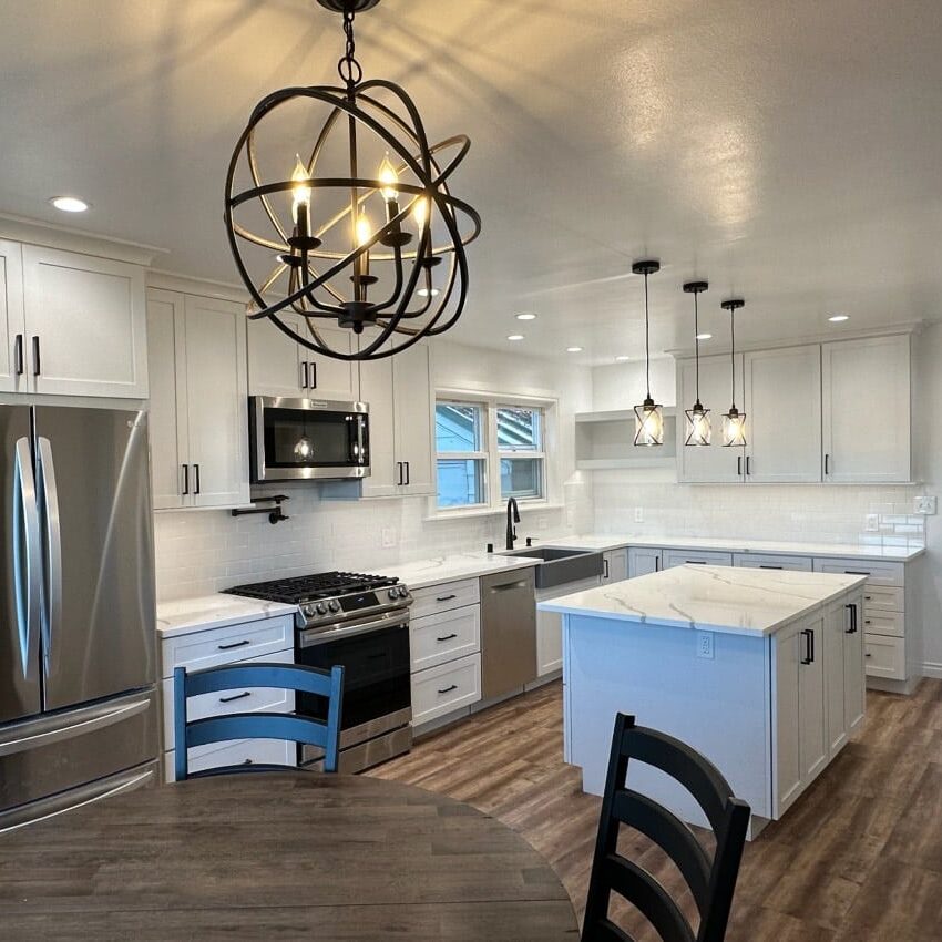 Modern kitchen with white cabinets, stainless steel appliances, marble island, pendant lights, wood flooring, and a round dining table with black chairs in the foreground—expertly crafted by a trusted West Seattle remodeling contractor.