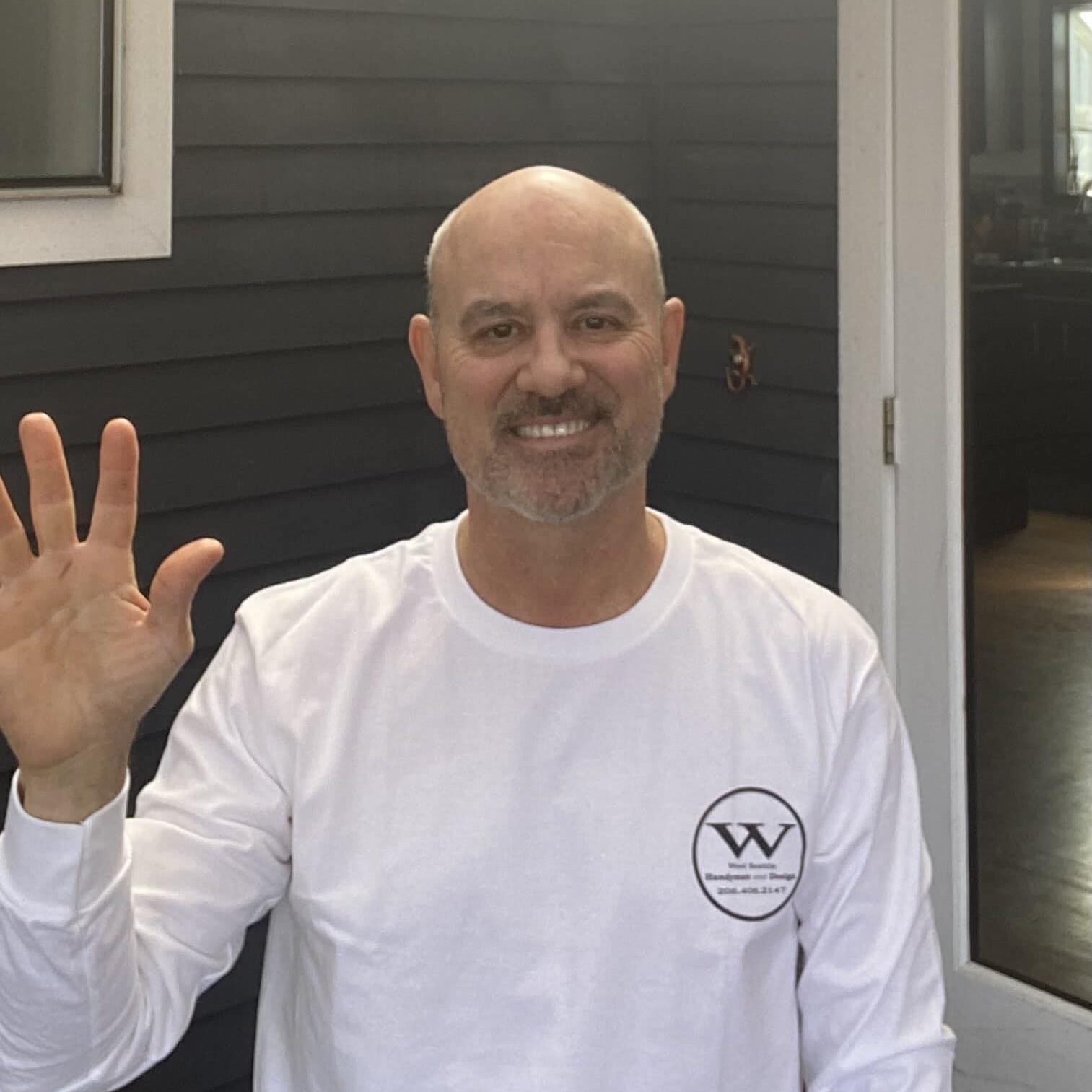 A man with a shaved head, goatee, and white shirt stands outdoors, smiling and waving at the camera in front of a dark-paneled wall and an open door—capturing the welcoming spirit of your trusted West Seattle remodeling contractor.