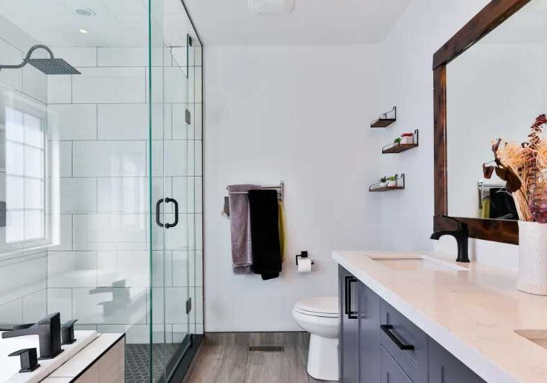 Modern bathroom with glass shower, a vanity with double sinks, large mirror, toilet, shelves, and a vase of dried flowers on the countertop—expertly designed by a trusted West Seattle remodeling contractor.